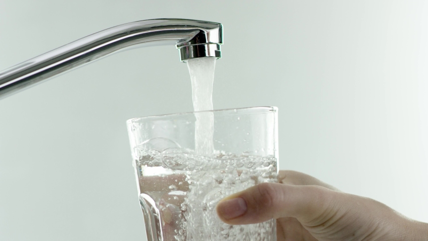 In the video, a water tap, water runs, hand brings a glass tumbler to the tap, the glass is completely filled with water, hand carries the glass, white background