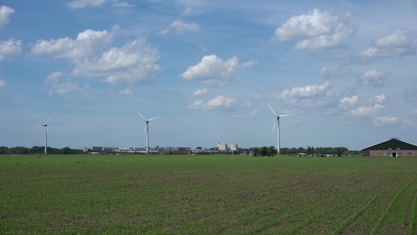 Low angle panoramic view of a wind farm with three wind turbines in Zoeterwoude in the Netherlands. Windswept grass in the foreground, buildings and clouds on blue sky in the background.