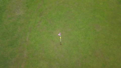 Drone shot of a red and white golf flag,ing in the wind, on a bright green golf field, on a sunny day