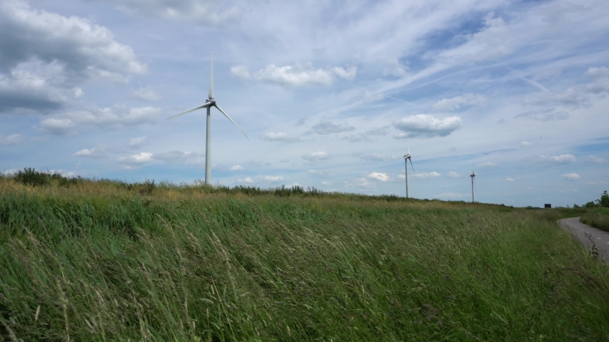 Low angle panoramic view of a wind farm with three wind turbines in rural environment in Zoeterwoude. Windswept grass in the foreground, clouds on blue sky in the background.