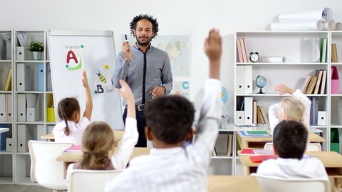Tracking shot of male Arab primary school teacher standing by whiteboard and teaching alphabet to little schoolchildren sitting at their desks and raising hands to answer question