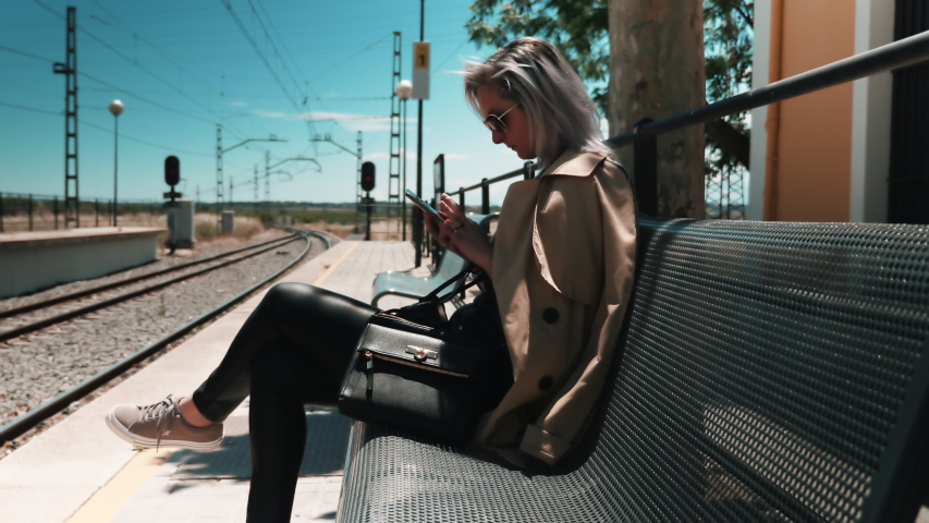 Young beautiful tourist woman checking her smartphone while waiting for the commuter train, wearing sunglasses on a bright sunny day, 4k side view