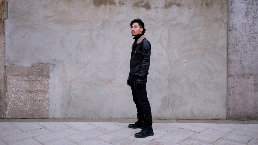 Chinese young mustached man dancing on a street next to an urban concrete wall. He