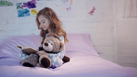 cute preteen child in pajamas sitting on bed with teddy bear and smiling while reading book