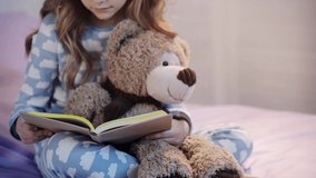 cropped view of preteen child in pajamas sitting on bed with teddy bear and reading book