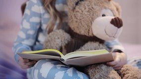 cropped view of preteen child in pajamas sitting on bed with teddy bear and reading book
