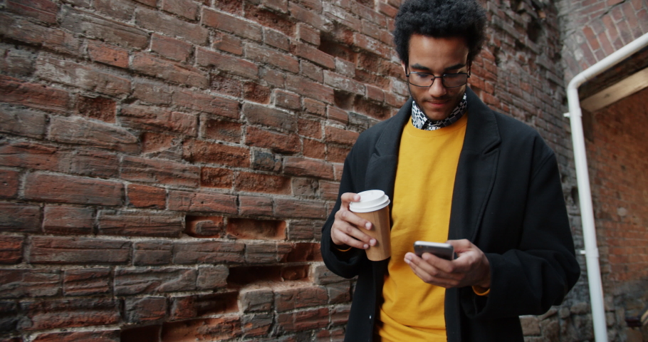 Slow motion of attractive African American guy using modern smartphone and holding to-go coffee walking outdoors near brick wall. People and technology concept.