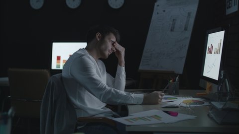 Unhappy businessman browsing business graphs and charts in night office. Sad analyst working overtime with financial reports in late office. Bankrupt and bad business statistic