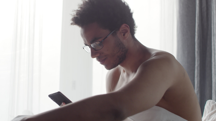 Medium shot of young black man waking up in the morning and using smartphone to browse internet | Shutterstock HD Video #1032390410