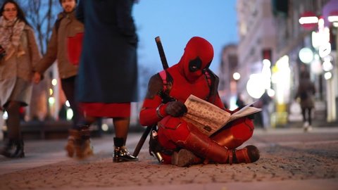 DNIPRO, UKRAINE - MARCH 28, 2019: Deadpool cosplayer sits on the sidewalk and reads newspaper in his hands on background of city street.