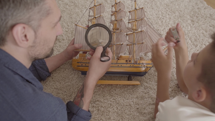 Father and his son lying at home on the floor on the fluffy carpet playing with ship model close-up. The man looking oh detail through a loupe. Family spending time together. Back view