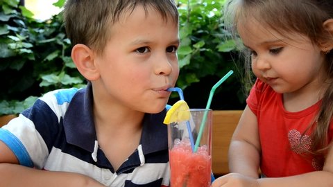 Close up of a smiling boy and girl drinking using two straws. sharing a smoothie sitting outdoors. cute brother and sister drink refreshing cocteil or watermelon smoothie from one glass against the