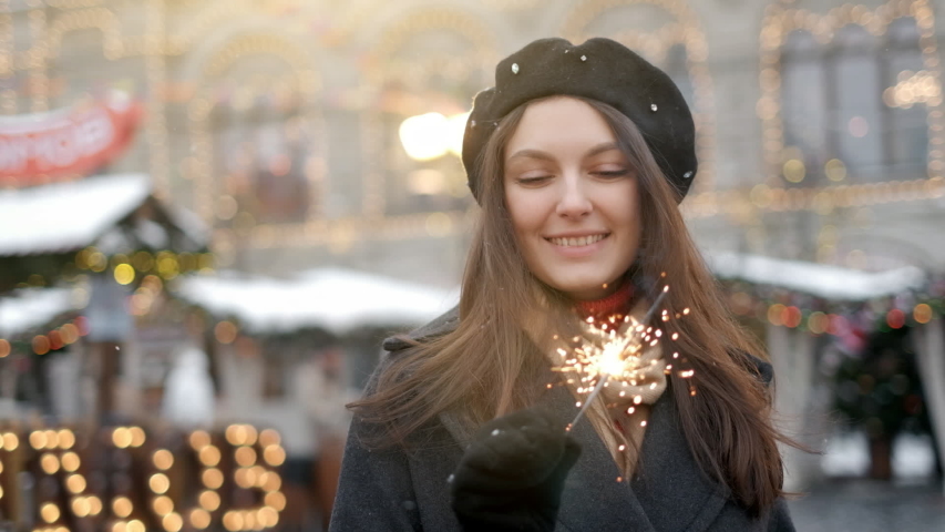 Outdoor of young beautiful happy smiling girl holding sparkler, walking on street. Model looking up, wearing stylish winter clothes. Waist up. Christmas, New Year, concept. Magic snowfall. Toned