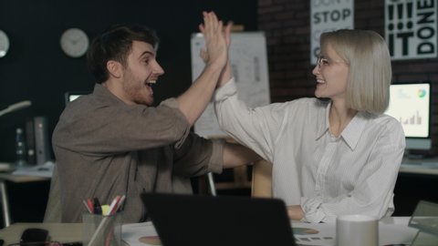 Happy business couple giving high five in dark office. Successful business partners winning good results in night office. Excited team celebrating business success.