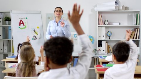 Medium shot of female primary teacher teaching alphabet to group of little schoolchildren raising their hands to answer questions