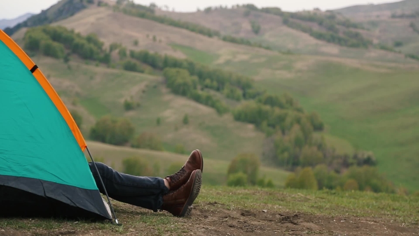 feet in brown boots protrude from a green tent with an orange stripe, on top of a mountain. in the background of the forest.