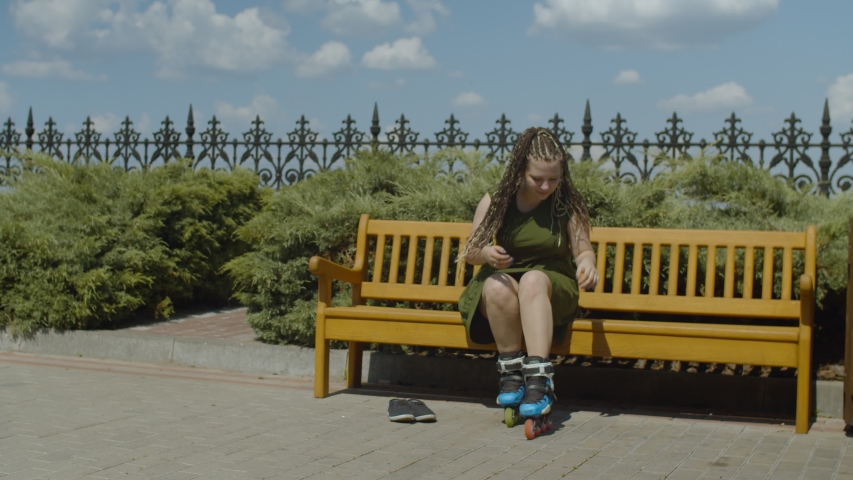 Stylish female with long ponytail from afro-braids unfastening and taking roller skates off while sitting on park bench after ride. Young woman roller going to relax after workout in public park.