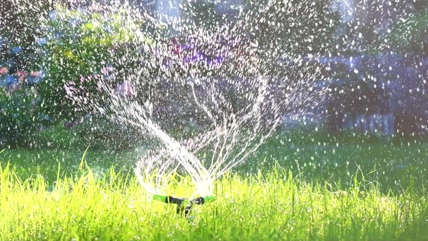 Garden Grass Watering. Smart garden activated with full automatic sprinkler irrigation system working in a green park, watering lawn, flowers and trees sprinkler head rotation. 4K UHD slow motion Royalty-Free Stock Footage #1032456548