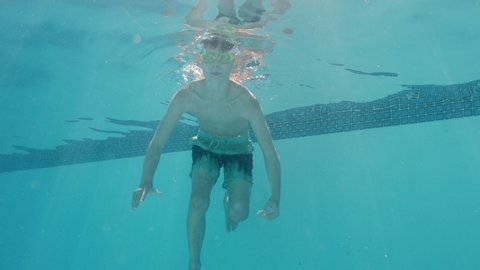 funny little boy swimming underwater in pool smiling waving hand enjoying swim in crystal clear water wearing goggles enjoying summer 4k