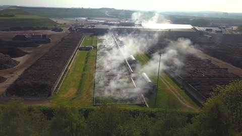 Panoramic drone shot of an industrial compost biofilter, with steam going up, surrounded by dunghills and green fields
