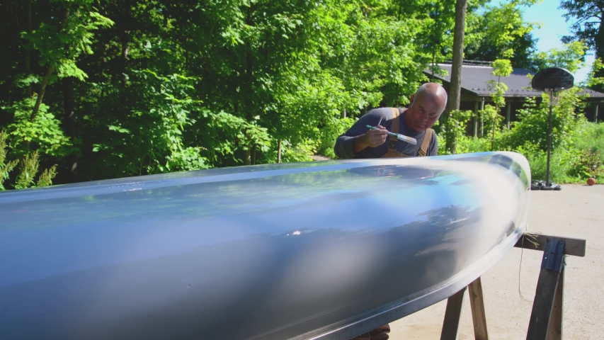 A mature man happily painting a canoe on a warm summer morning.  He is outside on his driveway.