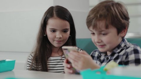 Two cheerful kids using a smartphone while eating a school lunch. Cute girl taking her phone away from a silly little boy.