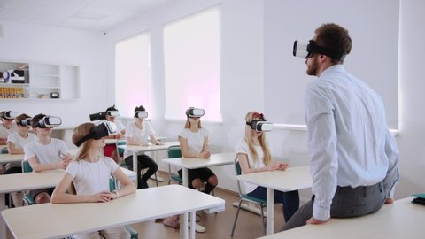 Computer science wwith virtual reality at a school lesson. Teacher explaining how future gadgets work while students with headsets listening carefully.