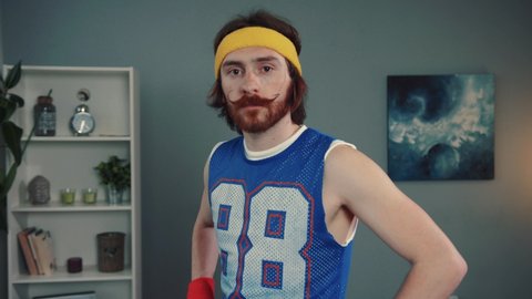 Portrait of amazed young bearded man hipster in retro sports outfit looking with a surprised expression standing in his living room at home.