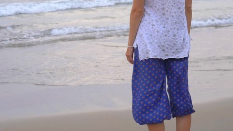 A young slim girl of European appearance with blond short hair, in blue wide trousers Aladdin and white T-shirt stands on the shore with his back to the viewer and looks at the sea in the evening.
