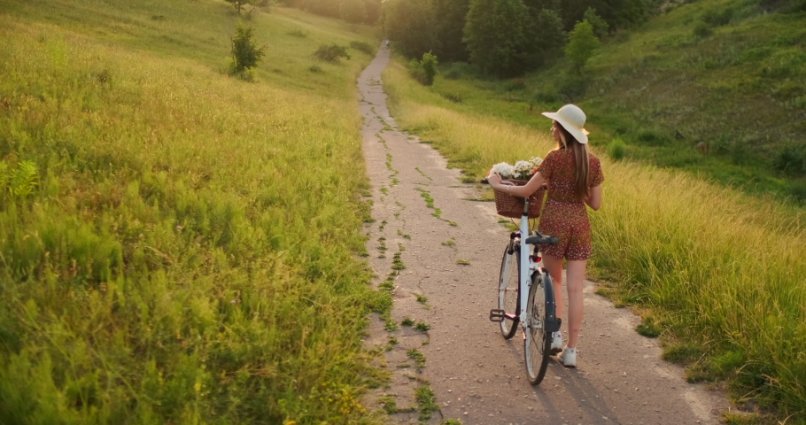 Back plan: Beautiful blonde in dress and retro bike walking on the road in the summer field.
