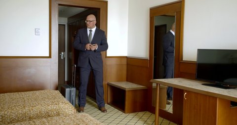 A bald man in glasses, business suit and brown shoes comes into the room. Puts his travel bag on wheels at the entrance, looks at himself in the mirror, straightens his tie