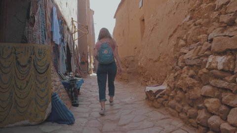 Following a blonde woman walking through a narrow street filled with market stalls displaying colorful scarfs and clothes in an outdoor market place in a Marrakech Souk in Morocco on a sunny day