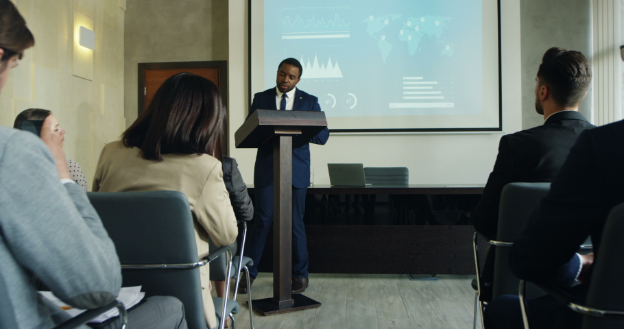 African American young good looking businessman demonstrating some graphics and charts on the big screen behind while speaking at the lecturn in front of the audience. | Shutterstock HD Video #1032512081