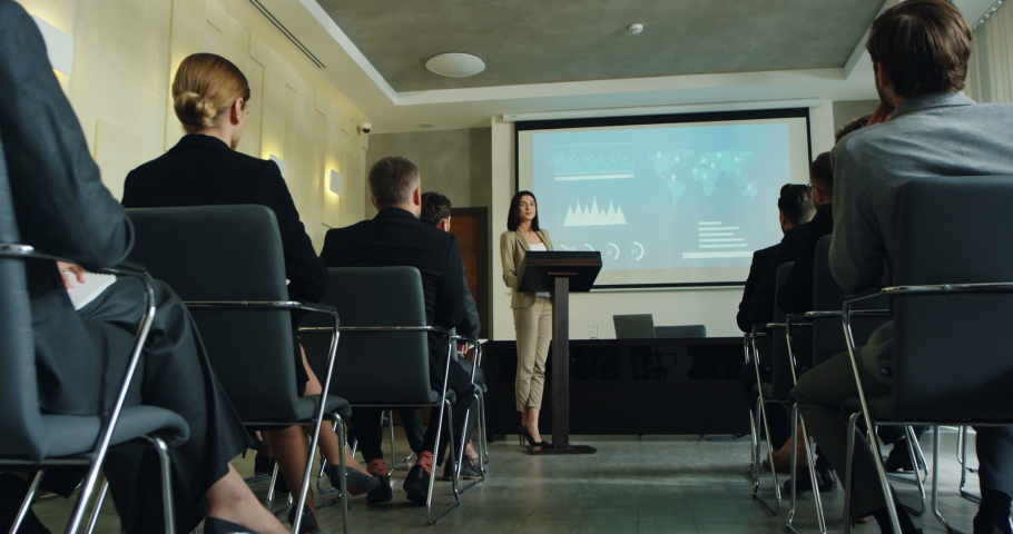 Caucasian young good looking brunette businesswoman demonstrating some graphics and charts on the big screen behind while speaking at the lecturn in front of the audience. | Shutterstock HD Video #1032512123
