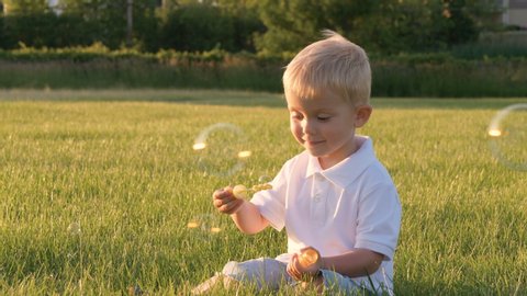 Cute little boy in white polo-shirt playing outdoors, blowing soap bubbles, having fun in a park. Summer, sunny