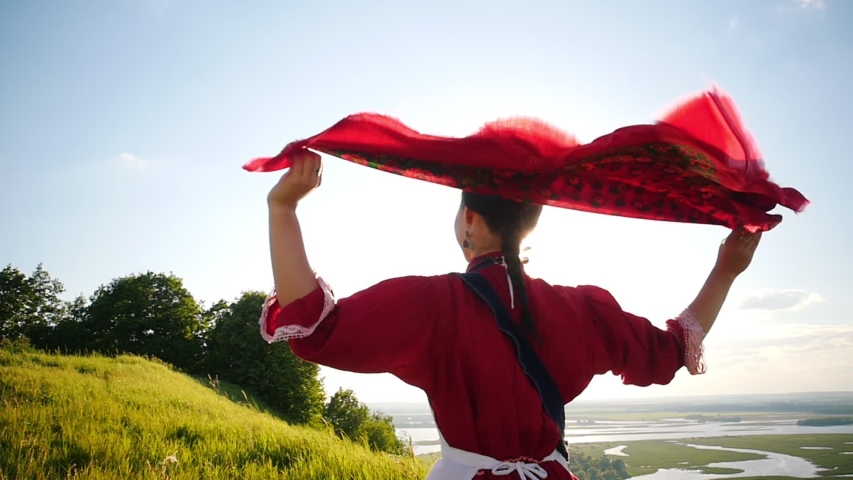 A young woman in russian traditional clothes walking on the green field - her red handkerchief flutters by the wind