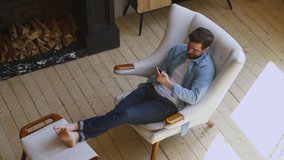 Happy young man sit on comfortable chair in modern country house room with fireplace relax hold smartphone using phone apps play mobile games lounge at cozy home on wooden floor, top view from above