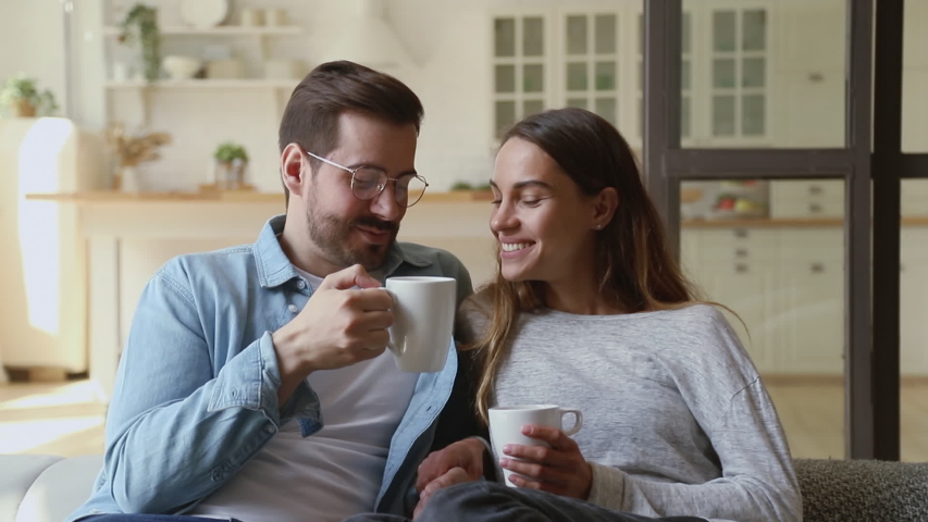 Happy young family couple relaxing talking laughing holding cups drinking coffee tea sitting on sofa together in living room, loving husband and wife bonding enjoying pleasant conversation at home Royalty-Free Stock Footage #1032517142