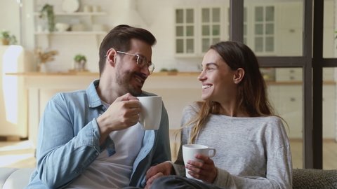 Happy young family couple relaxing talking laughing holding cups drinking coffee tea sitting on sofa together in living room, loving husband and wife bonding enjoying pleasant conversation at home