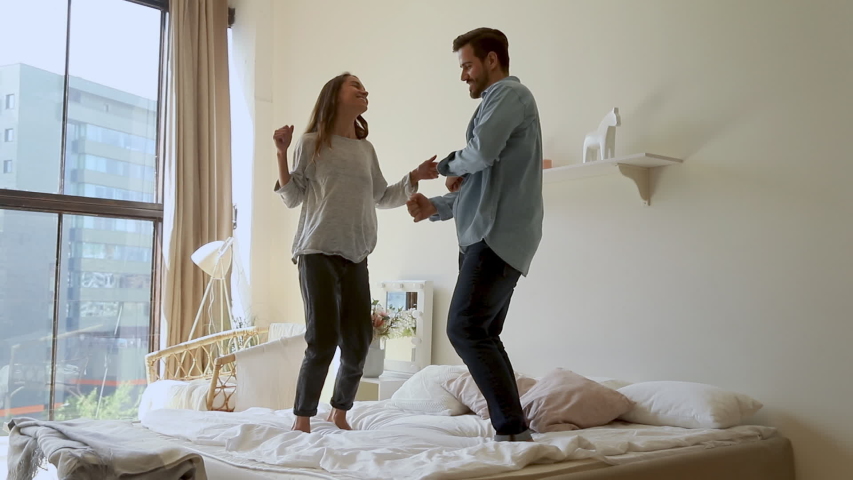 Happy funny young family couple dancing jumping on bed mattress, active carefree husband and wife having fun laughing enjoying honeymoon party together in morning in cozy bedroom interior at home Royalty-Free Stock Footage #1032517148