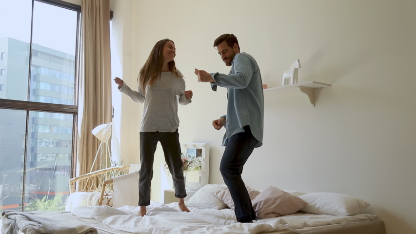 Happy funny young family couple dancing jumping on bed mattress, active carefree husband and wife having fun laughing enjoying honeymoon party together in morning in cozy bedroom interior at home Royalty-Free Stock Footage #1032517148