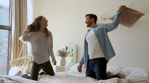Happy young romantic family couple playing pillow fight on bed, cheerful carefree husband and wife having fun laughing in bedroom interior in morning enjoy lifestyle activity game together at home