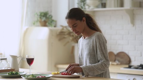 Happy young woman holding knife cutting fresh organic vegetables for salad in kitchen, smiling fit vegetarian girl prepare healthy dinner make surprise for husband enjoy glass of wine cooking at home
