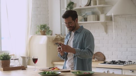 Happy young man preparing romantic surprise using smartphone mobile apps, smiling husband cooking healthy food cutting salad for family dinner in kitchen holding phone search vegan recipe at home