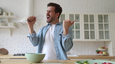 Happy funny young man holding beater microphone singing song dancing listening to music in kitchen, funky carefree guy preparing morning breakfast meal cooking healthy food having fun alone at home