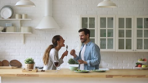 Funny young couple laughing holding kitchenware singing song together dancing to music enjoying cooking in modern cozy kitchen, happy carefree husband and wife having fun preparing food at home