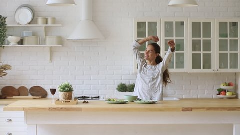 Carefree happy young woman housewife dancing alone cooking meal in modern kitchen, funny cheerful active pretty girl preparing food healthy dinner vegetable salad at home having fun listening music