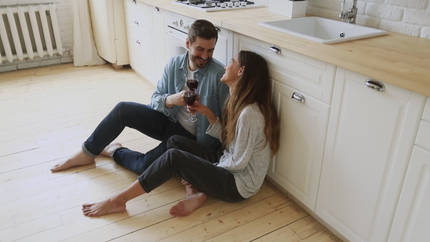Affectionate young family couple happy husband and wife sit on cozy kitchen room floor hold glasses drink red wine talk laugh celebrate holiday new home purchase rent mortgage investment, above view