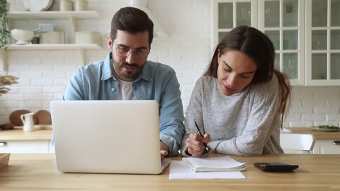 Young family couple husband and wife doing paperwork planning budget expenses discussing money finances payments calculating paying bills using laptop computer together sit at kitchen table at home