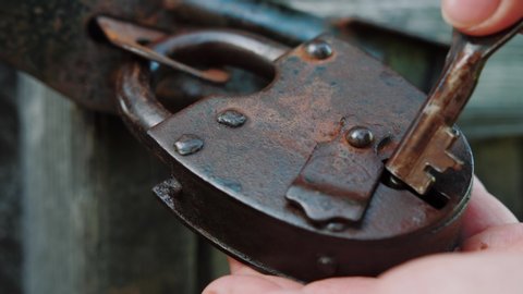 Old vintage rusty iron lock on retro wooden door. Female hand open padlock with key. Woman’s hands turn key in keyhole. Close up, slow motion.